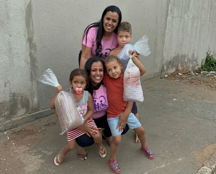 Duas voluntárias do Cidadania que Acolhe, de camiseta rosa, sorrindo abraçadas a quatro crianças que seguram sacolas de doação