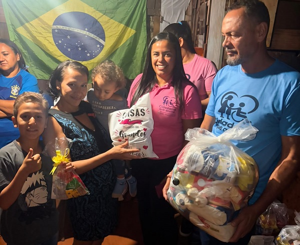 Equipe do Instituto Cidadania que Acolhe entregando cestas e kits a uma família, em frente a uma bandeira do Brasil